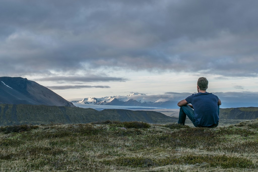 Person sitting on a grassy ridge, looking out at snow-capped mountains and a glacier under a cloudy sky