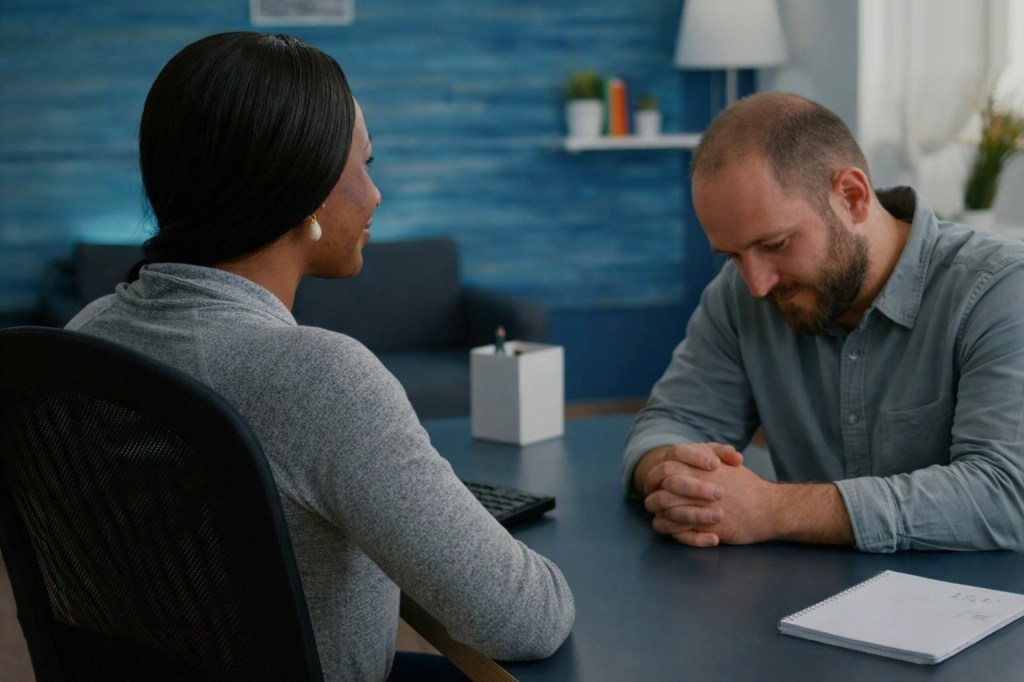 Manager listening kindly to an employee who sits with his head bowed and hands clasped