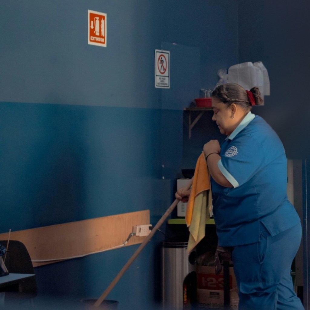 Housekeeper in a blue uniform carrying a mop and cleaning cloths through a dimly lit workspace