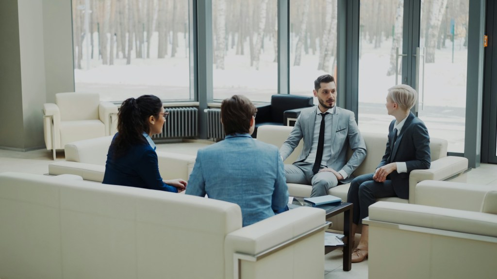 Business colleagues in conversation on white sofas in a bright, glass-walled office lobby